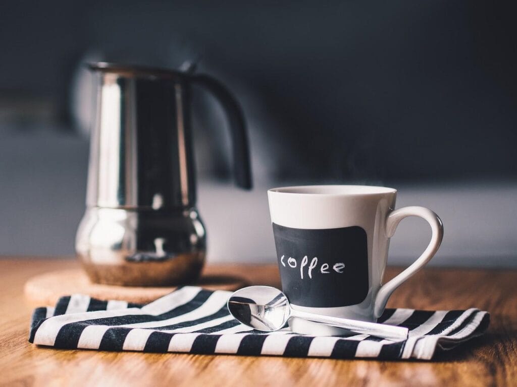 A cup of coffee on a napkin with a spoon beside it, next to a coffee pot, illustrating how to make coffee from beans.
