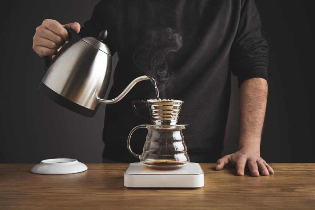 A person pouring hot water from a gooseneck kettle into a pour-over coffee maker with steam rising.