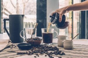 Pouring coffee into a glass mug surrounded by beans and cups illustrates can you brew coffee with whole beans